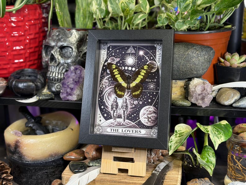 Puede incluir: Una impresi&oacute;n enmarcada en blanco y negro de la carta del tarot "Los Amantes", con una mariposa con alas amarillas y negras. La impresi&oacute;n se muestra en un caballete de madera, rodeada de plantas y objetos decorativos.
