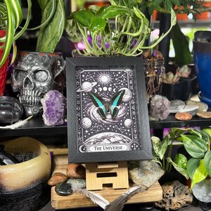Op de afbeelding: Een zwart-wit print in een lijst met een vlinder met turquoise en zwarte vleugels, tegen een hemelse achtergrond met planeten en sterren. De print is getiteld "The Universe". De afbeelding wordt weergegeven op een houten ezel, omringd door planten en decoratieve items.