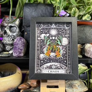 May include: A framed black and white print depicting a butterfly with orange and green wings, set against a celestial background with planets and stars. The word "CHAOS" is printed at the bottom. The frame is displayed on a wooden stand.
