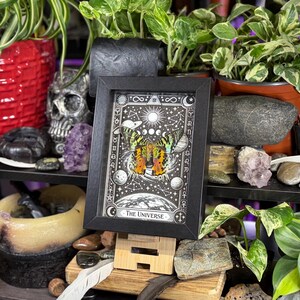 May include: A framed print of "The Universe" tarot card, featuring a colorful butterfly design with celestial elements. The black frame stands on a wooden easel, surrounded by plants, crystals, and decorative items. The artwork is in a black frame.