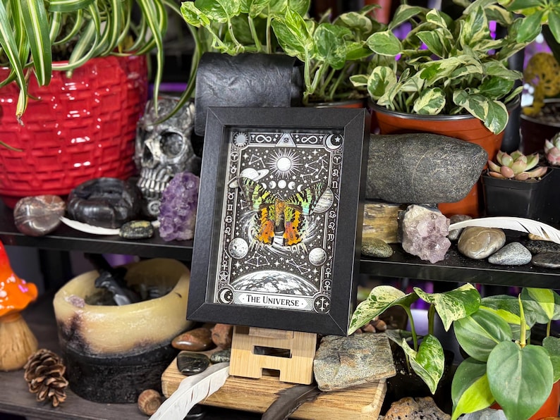 May include: A framed print of "The Universe" tarot card, featuring a colorful butterfly design with celestial elements. The black frame stands on a wooden easel, surrounded by plants, crystals, and decorative items. The artwork is in a black frame.