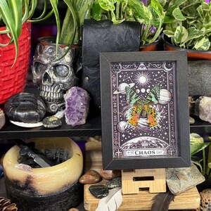 May include: A framed artwork with a black border, featuring a celestial design with a butterfly and the word "CHAOS." The artwork is displayed on a wooden stand, surrounded by plants, crystals, and decorative items. The color palette includes black, white, purple, and green.