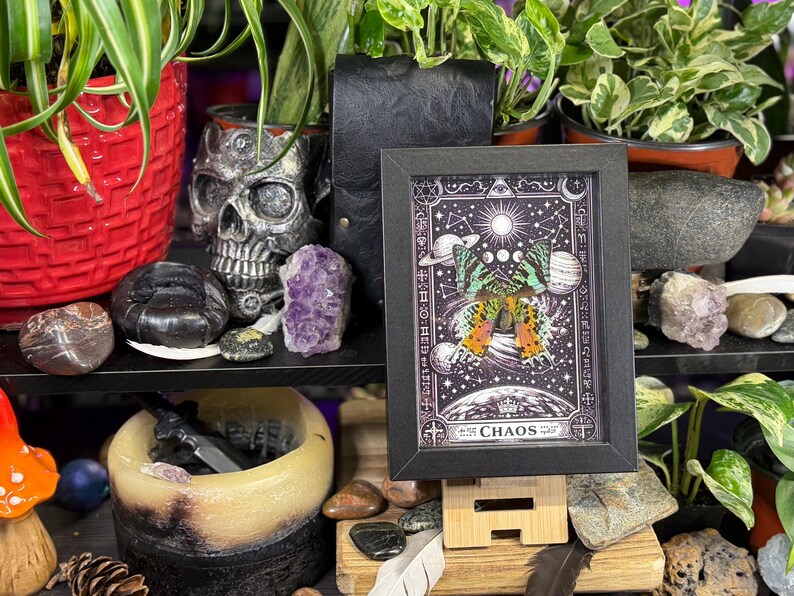 May include: A framed artwork with a black border, featuring a celestial design with a butterfly and the word "CHAOS." The artwork is displayed on a wooden stand, surrounded by plants, crystals, and decorative items. The color palette includes black, white, purple, and green.
