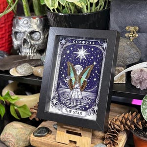 Op de afbeelding: Een ingelijst kunstwerk met een hemels ontwerp, met een figuur met vlindervleugels tegen een donkerblauwe sterrenachtergrond. De woorden "THE STAR" staan onderaan. De lijst is zwart en het kunstwerk staat op een houten standaard.