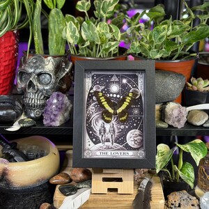 Puede incluir: Impresi&oacute;n enmarcada de una carta del tarot con una mariposa de alas amarillas y negras sobre un fondo celestial. La carta, titulada "The Lovers", se exhibe sobre un soporte de madera, rodeada de plantas y cristales.
