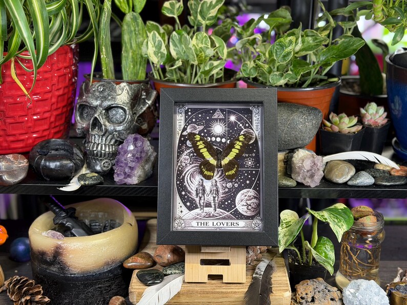 Puede incluir: Impresi&oacute;n enmarcada de una carta del tarot con una mariposa de alas amarillas y negras sobre un fondo celestial. La carta, titulada "The Lovers", se exhibe sobre un soporte de madera, rodeada de plantas y cristales.