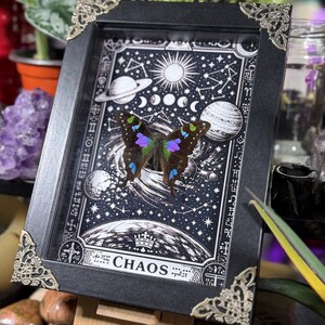Może przedstawiać: Oprawiona grafika przedstawiająca motyla z niebieskimi, fioletowymi i zielonymi skrzydłami, na tle nieba z planetami, gwiazdami i napisem "CHAOS". Czarna rama ma ozdobne narożniki.