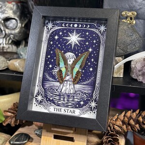 Op de afbeelding: Een ingelijst kunstwerk met een vlinder met blauwe en bruine vleugels tegen een sterrenhemel. Het kunstwerk is getiteld "THE STAR" en maakt deel uit van een tarotkaartthema. Het zwarte frame wordt gepresenteerd op een houten standaard.