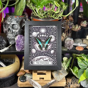 Op de afbeelding: Een ingelijste zwart-wit print met een vlinder met turquoise en zwarte vleugels, tegen een hemelse achtergrond met planeten, sterren en de tekst "The Universe". De print wordt op een houten ezel getoond.
