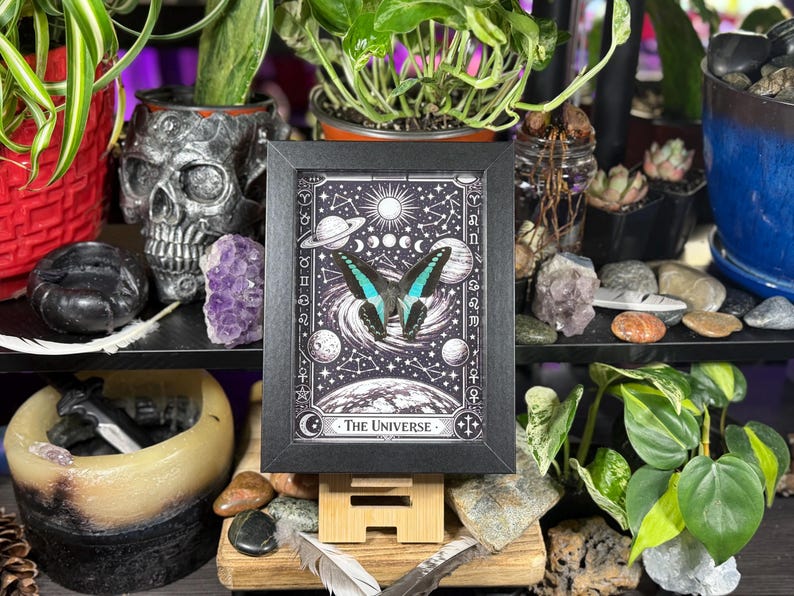 Op de afbeelding: Een ingelijste zwart-wit print met een vlinder met turquoise en zwarte vleugels, tegen een hemelse achtergrond met planeten, sterren en de tekst "The Universe". De print wordt op een houten ezel getoond.