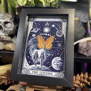 May include: A framed art print with a celestial theme, depicting a butterfly and two astronauts. The artwork is mainly blue and white, with orange and yellow details. The text "THE LOVERS" is at the bottom. The frame is black.