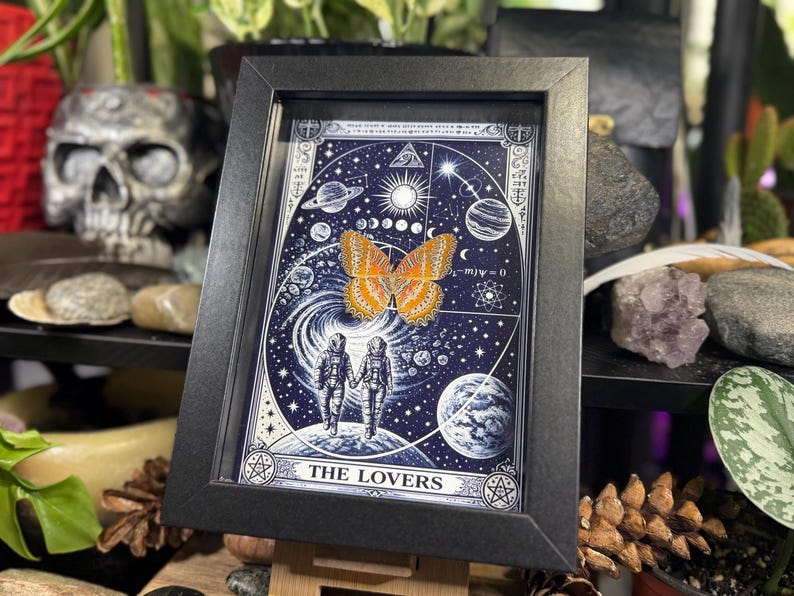 May include: A framed art print with a celestial theme, depicting a butterfly and two astronauts. The artwork is mainly blue and white, with orange and yellow details. The text "THE LOVERS" is at the bottom. The frame is black.