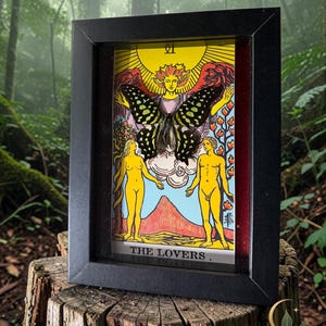 May include: A black rectangular shadow box frame containing a tarot card titled THE LOVERS. The card features a yellow sun, a central figure, and two standing figures. A dark butterfly with light green spots is mounted over the centre of the card.