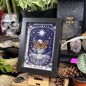 May include: Framed art print featuring a celestial illustration. The artwork depicts a figure holding a butterfly with orange and brown wings, set against a starry night sky. The words "THE STAR" are at the bottom of the print.