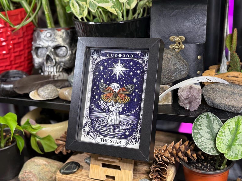 May include: Framed art print featuring a celestial illustration. The artwork depicts a figure holding a butterfly with orange and brown wings, set against a starry night sky. The words "THE STAR" are at the bottom of the print.
