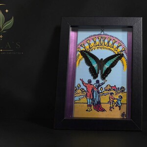 Puede incluir: Una obra de arte enmarcada con una mariposa negra y azul verdoso montada sobre una ilustraci&oacute;n de la carta del tarot del Diez de Copas. La imagen muestra a una familia mirando hacia un arco&iacute;ris de diez copas doradas en un cielo azul, dentro de un marco negro.
