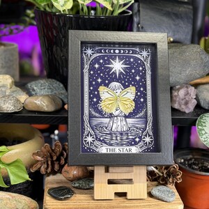 May include: A framed print of "The Star" tarot card. The artwork depicts a figure holding a yellow butterfly against a starry night sky. The black framed print is displayed on a small wooden easel.