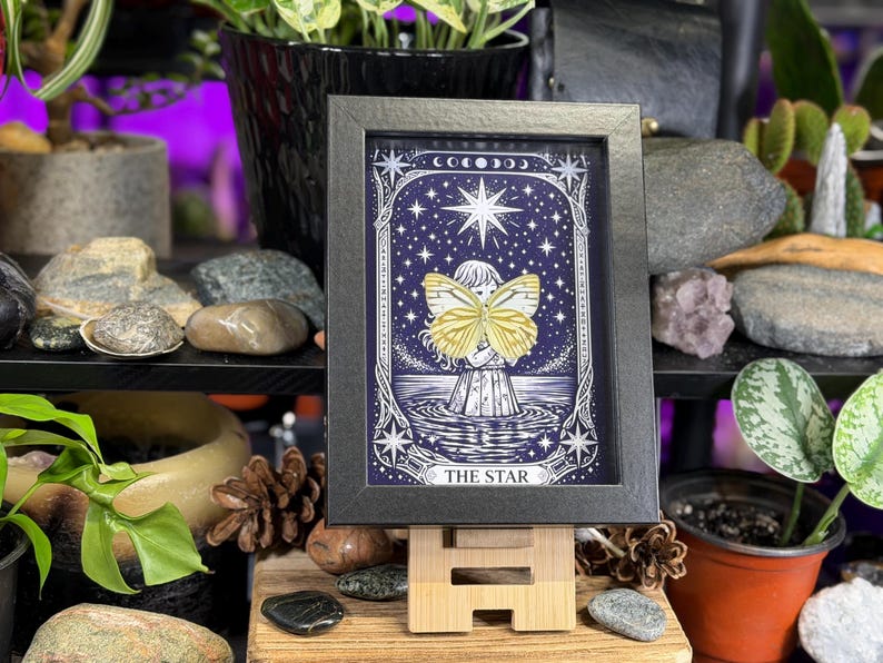 May include: A framed print of "The Star" tarot card. The artwork depicts a figure holding a yellow butterfly against a starry night sky. The black framed print is displayed on a small wooden easel.