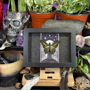 May include: A framed artwork depicting a butterfly with yellow and black wings against a starry night sky. The black-framed piece is displayed on a wooden stand. The background includes plants, crystals, and decorative items.