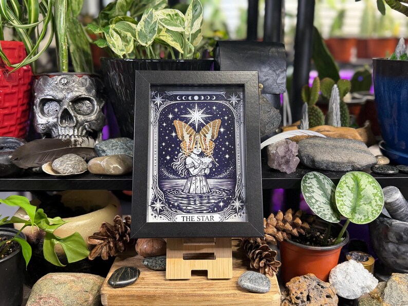 May include: A framed print of "The Star" tarot card, depicting a woman with butterfly wings against a starry night sky. The black frame is on a wooden easel, surrounded by plants, rocks, and a skull.