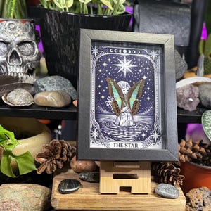 Op de afbeelding: Een ingelijste tarotkaartafdruk van "The Star" met een figuur met vlindervleugels tegen een sterrenhemel. De zwarte lijst staat op een houten standaard, omringd door planten en decoratieve items.