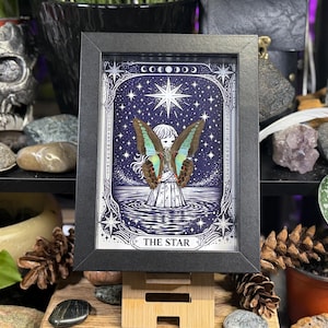 Op de afbeelding: Ingelijste illustratie met een hemels ontwerp met een centrale figuur met vlindervleugels, tegen een sterrenhemel. Het kunstwerk is onderaan voorzien van het opschrift "THE STAR", is zwart omlijst en staat op een houten standaard.