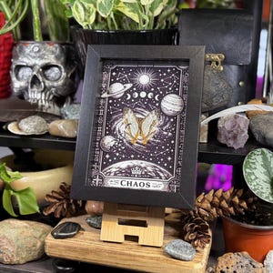 May include: A framed print with a black border, featuring a celestial design with planets, stars, and a butterfly. The word "CHAOS" is at the bottom. The print is displayed on a wooden easel.