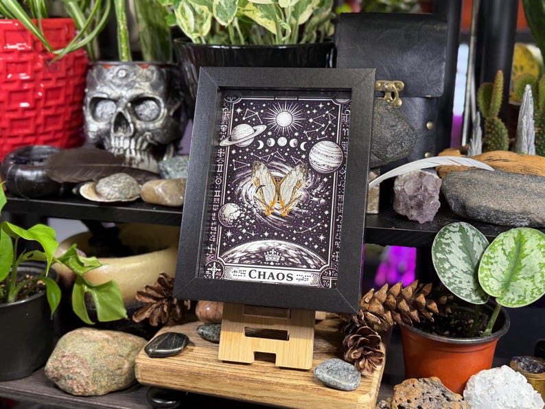 May include: A framed print with a black border, featuring a celestial design with planets, stars, and a butterfly. The word "CHAOS" is at the bottom. The print is displayed on a wooden easel.