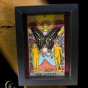 May include: A black rectangular shadow box frame containing a tarot card titled THE LOVERS with the Roman numeral VI. A large, dark butterfly with light green spots is mounted over the centre of the card, which depicts two figures.
