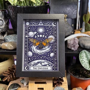 K&ouml;nnte beinhalten: Ein gerahmter Druck mit einem Himmelsdesign und einer Mottenillustration. Das Kunstwerk ist vor einem dunkelblauen Hintergrund mit wei&szlig;en Sternen, Planeten und dem Text "The Universe" platziert. Der Rahmen ist schwarz.