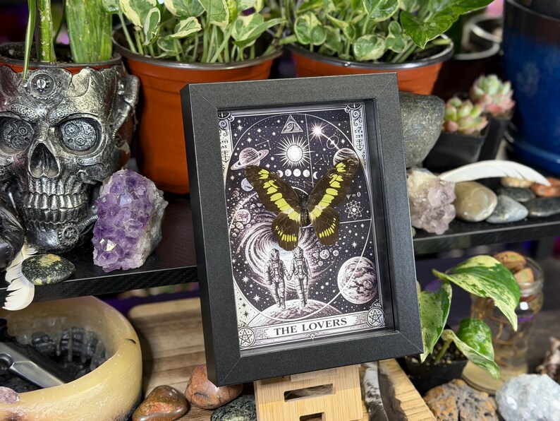 Puede incluir: Una impresi&oacute;n enmarcada de una mariposa con alas amarillas y negras, sobre un fondo celestial con la inscripci&oacute;n "THE LOVERS". El marco negro se exhibe sobre un soporte de madera, rodeado de plantas, cristales y objetos decorativos.