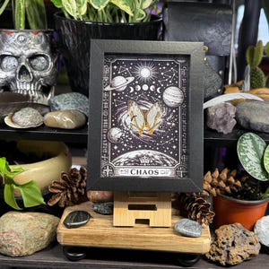 May include: A framed art print with a celestial theme, showing planets, stars, and a butterfly. The print is in a black frame, displayed on a wooden easel. The word "CHAOS" is at the bottom of the print.