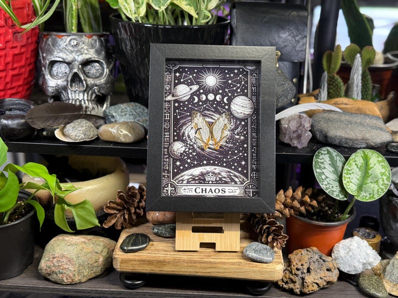 May include: A framed art print with a celestial theme, showing planets, stars, and a butterfly. The print is in a black frame, displayed on a wooden easel. The word "CHAOS" is at the bottom of the print.