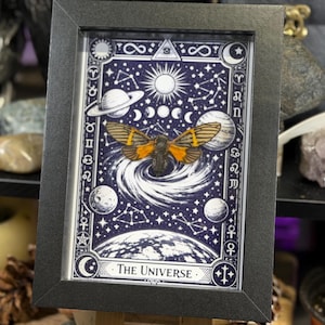 K&ouml;nnte beinhalten: Gerahmtes Kunstwerk mit einem himmlischen Design, das eine Motte im Zentrum zeigt. Das Design hat einen dunkelblauen Hintergrund mit wei&szlig;en Sternen, Planeten und der Sonne. Der Text "The Universe" steht unten. Der Rahmen ist schwarz.