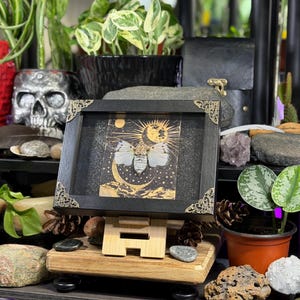 May include: A framed artwork with a celestial theme, depicting a moth, sun, and moon. The black frame has decorative corners and is displayed on a wooden easel. The artwork is surrounded by plants and decorative objects.