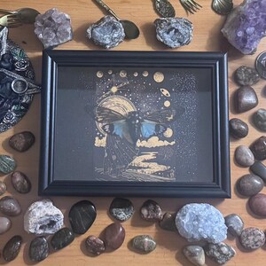 May include: A black framed artwork with a celestial moth design, surrounded by crystals, stones, and decorative metal pieces. The artwork has a dark background with gold accents. The image is set on a wooden surface.