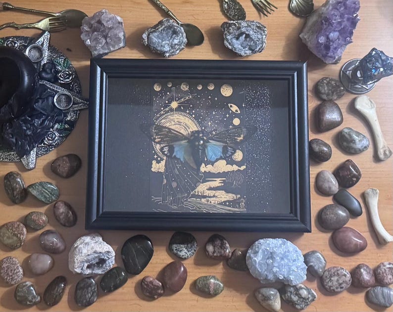 May include: A black framed artwork with a celestial moth design, surrounded by crystals, stones, and decorative metal pieces. The artwork has a dark background with gold accents. The image is set on a wooden surface.