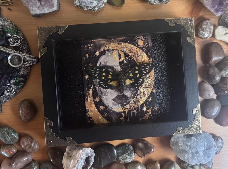 May include: A black framed art piece featuring a moth with yellow and white markings, set against a celestial background of a moon and stars. The artwork is surrounded by decorative corner accents and sits on a wooden surface with various stones.