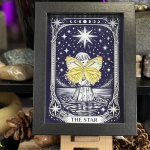 May include: Framed artwork depicting a celestial scene with a central figure and a large yellow butterfly. The artwork is set against a dark blue starry background, with the words "THE STAR" at the bottom. The frame is black.