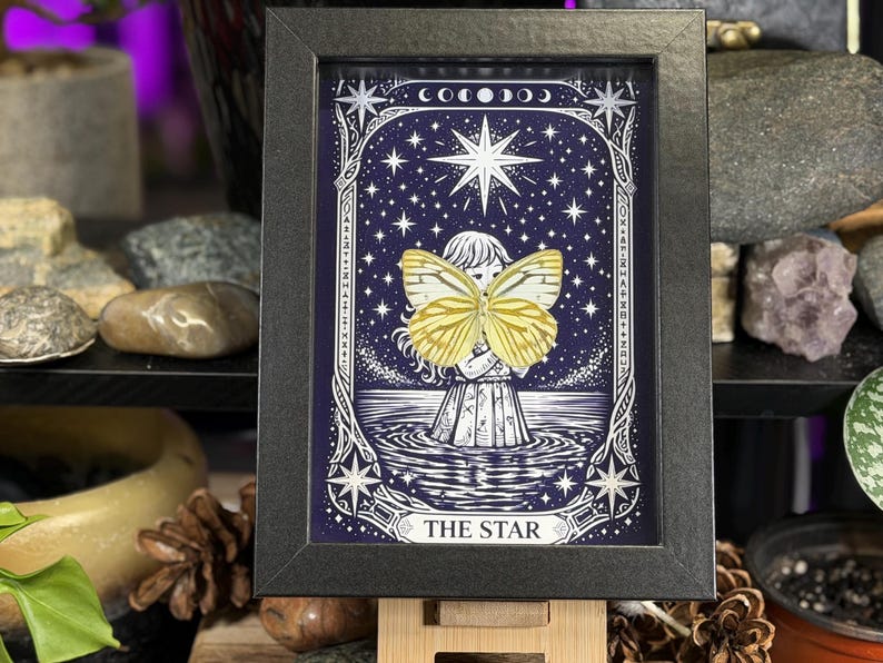 May include: Framed artwork depicting a celestial scene with a central figure and a large yellow butterfly. The artwork is set against a dark blue starry background, with the words "THE STAR" at the bottom. The frame is black.