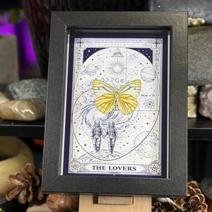 K&ouml;nnte beinhalten: Gerahmtes Kunstwerk mit einem gelben Schmetterling &uuml;ber einer Himmelsillustration mit zwei Astronauten, die sich an den H&auml;nden halten. Das Kunstwerk ist in einem schwarzen rechteckigen Rahmen gerahmt und der Text "THE LOVERS" befindet sich unten.