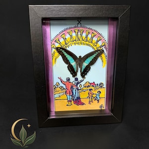 Puede incluir: Una obra de arte enmarcada que presenta una ilustraci&oacute;n de la carta del tarot Diez de Copas con un esp&eacute;cimen de mariposa central. La mariposa tiene alas de color marr&oacute;n oscuro con marcas azul turquesa. La carta muestra a una familia bajo un arco&iacute;ris de diez copas doradas.