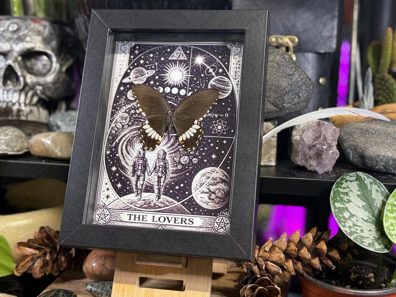 Puede incluir: Una impresi&oacute;n en blanco y negro enmarcada con una mariposa y un dise&ntilde;o de carta del tarot. La impresi&oacute;n incluye las palabras "THE LOVERS" en la parte inferior. La mariposa tiene alas oscuras con marcas blancas. El marco se exhibe en un caballete de madera.