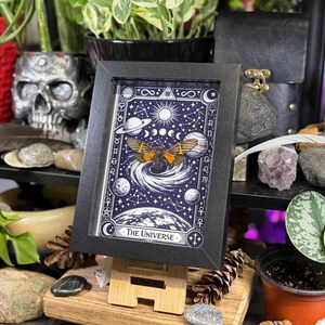 K&ouml;nnte beinhalten: Ein gerahmter Druck der Tarotkarte "Das Universum". Das Kunstwerk zeigt einen Schmetterling mit orangefarbenen und schwarzen Fl&uuml;geln, umgeben von Himmelsk&ouml;rpern auf einem dunkelblauen Hintergrund. Der Rahmen ist schwarz und der Druck wird auf einer Holzstaffelei ausgestellt.