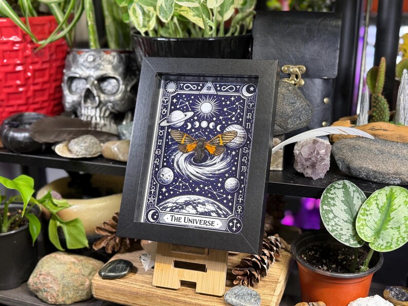 K&ouml;nnte beinhalten: Ein gerahmter Druck der Tarotkarte "Das Universum". Das Kunstwerk zeigt einen Schmetterling mit orangefarbenen und schwarzen Fl&uuml;geln, umgeben von Himmelsk&ouml;rpern auf einem dunkelblauen Hintergrund. Der Rahmen ist schwarz und der Druck wird auf einer Holzstaffelei ausgestellt.