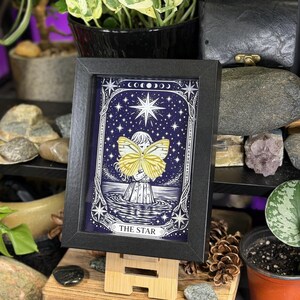 May include: A framed print of "The Star" tarot card. The artwork depicts a figure with yellow butterfly wings, standing in water under a starry night sky. The black frame holds the print, which is displayed on a small wooden easel. The overall theme is mystical and spiritual.
