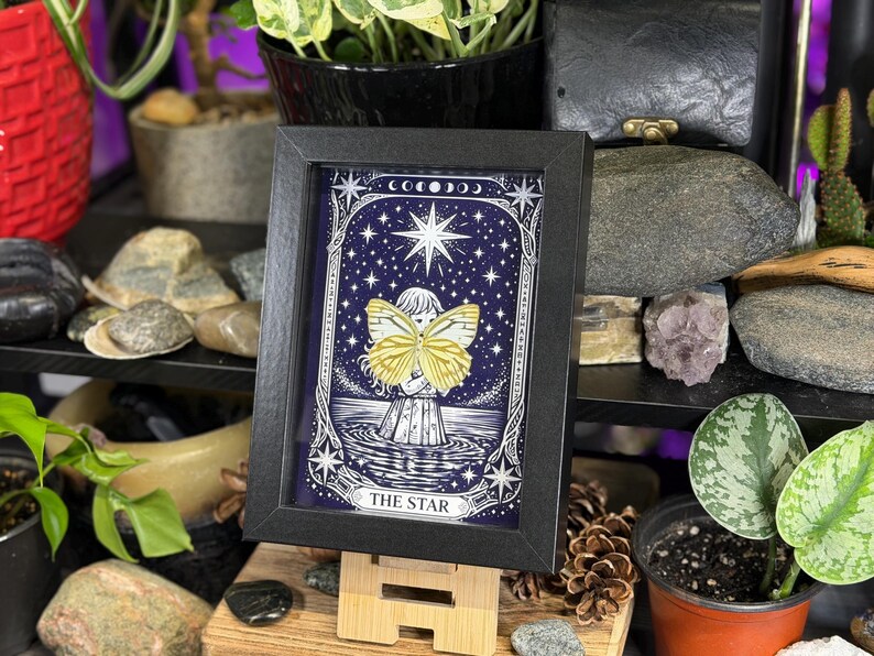 May include: A framed print of "The Star" tarot card. The artwork depicts a figure with yellow butterfly wings, standing in water under a starry night sky. The black frame holds the print, which is displayed on a small wooden easel. The overall theme is mystical and spiritual.