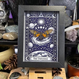 K&ouml;nnte beinhalten: Gerahmter Kunstdruck mit einem Himmelsmotiv, das Planeten, Sterne und eine Motte zeigt. Das Kunstwerk ist &uuml;berwiegend blau und wei&szlig;, mit orangefarbenen und schwarzen Akzenten auf der Motte. Der Text "The Universe" befindet sich unten.