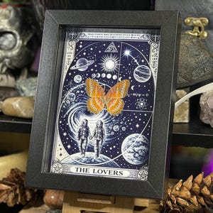May include: A framed art print with a celestial theme, showcasing a butterfly and two figures holding hands. The artwork is set against a dark blue backdrop with planets, stars, and the words "THE LOVERS" at the bottom.