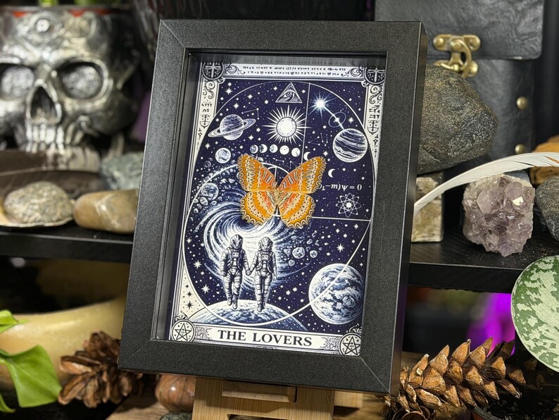 May include: A framed art print with a celestial theme, showcasing a butterfly and two figures holding hands. The artwork is set against a dark blue backdrop with planets, stars, and the words "THE LOVERS" at the bottom.